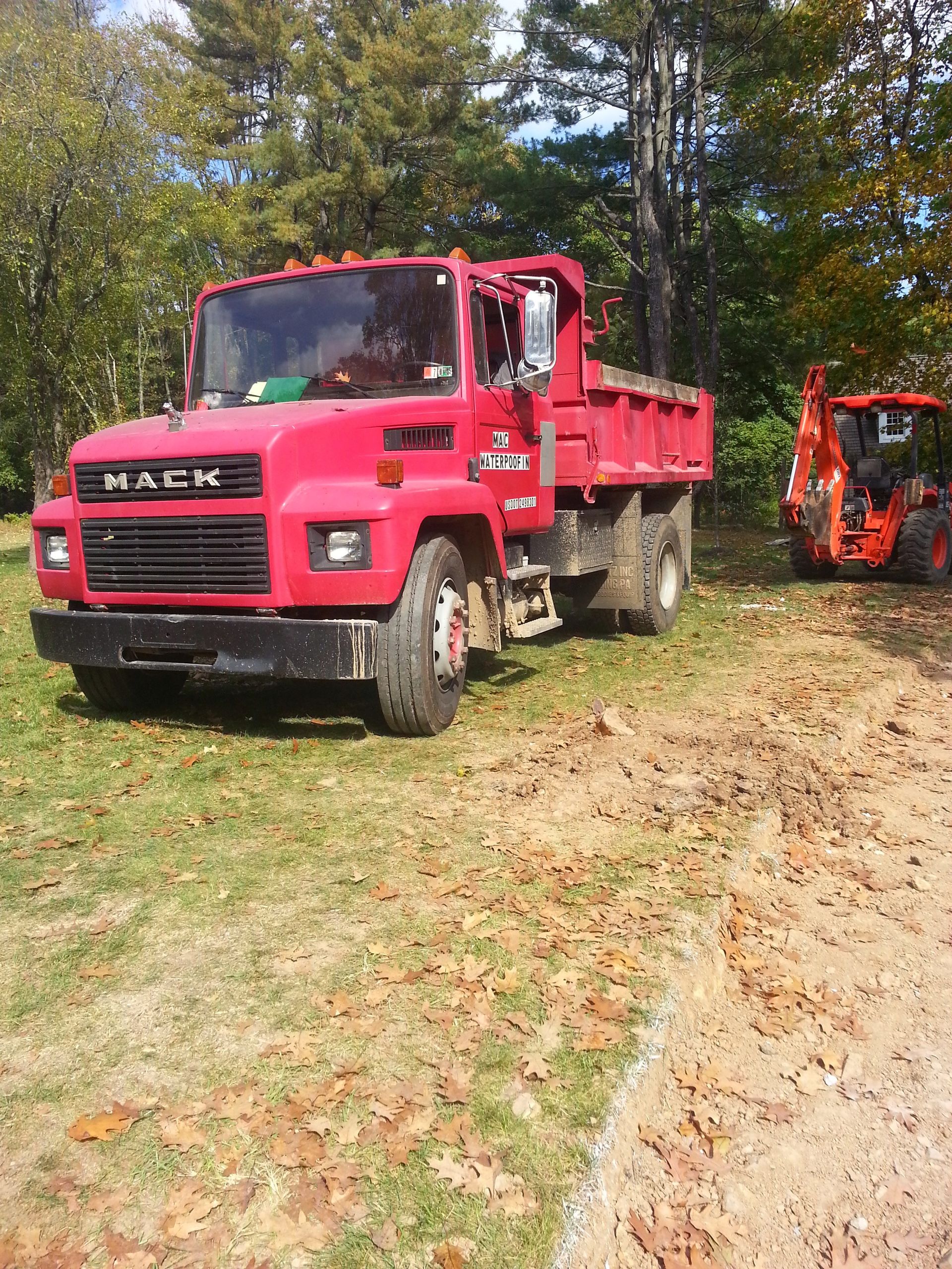 Red Mack dump truck on a grassy path with an orange excavator in the background near trees.
