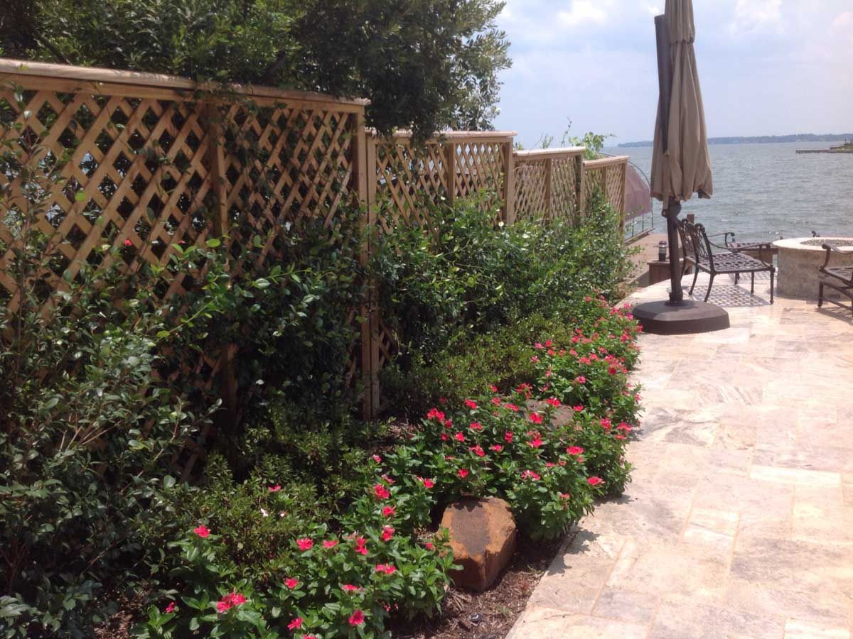 Wooden lattice fence with green shrubs and red flowers along a stone patio, overlooking a lake.