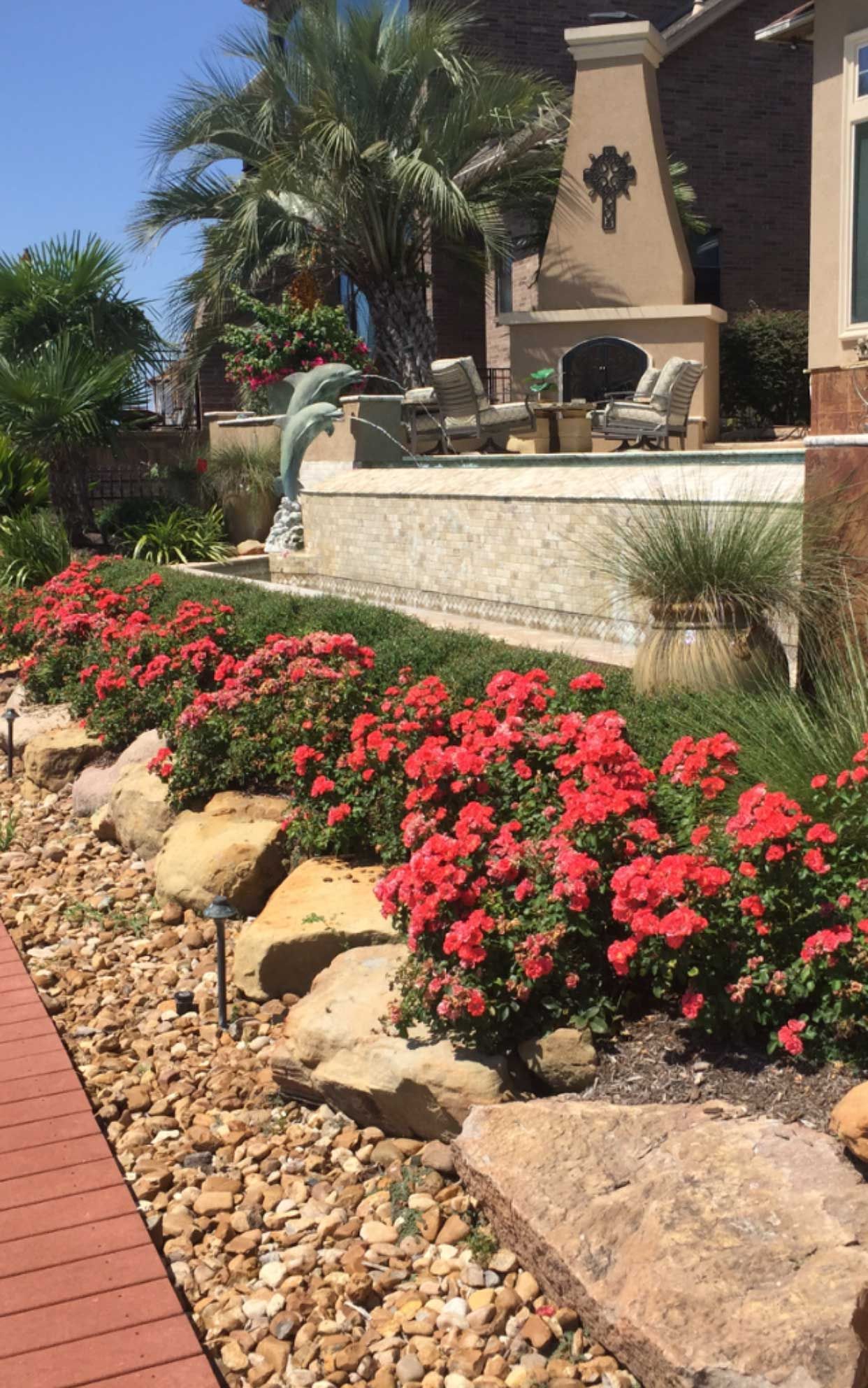 Red flowering bushes line a stone-edged garden path, leading to a patio with a fireplace and pool.