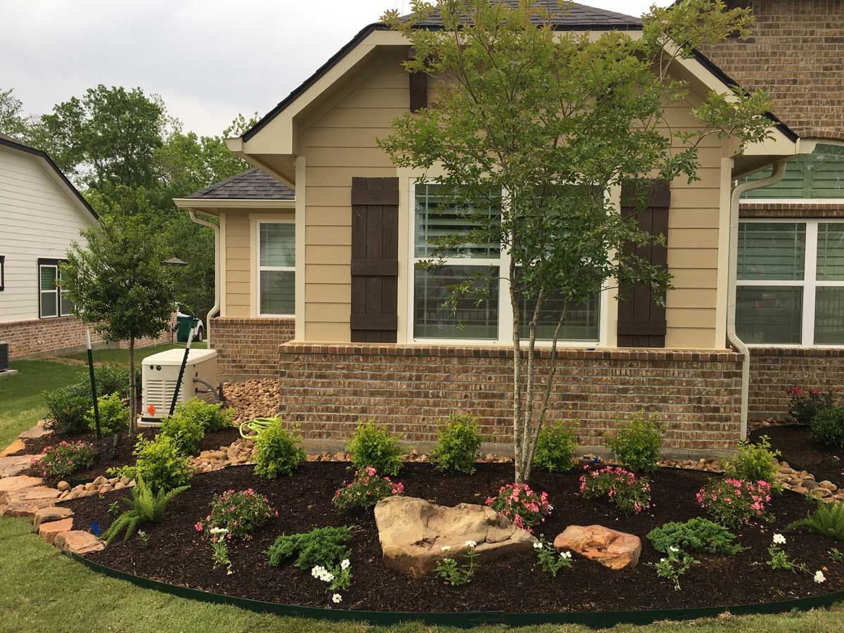 House exterior with landscaped flower bed and a tree. The house is beige with a brick base, and has brown shutters.