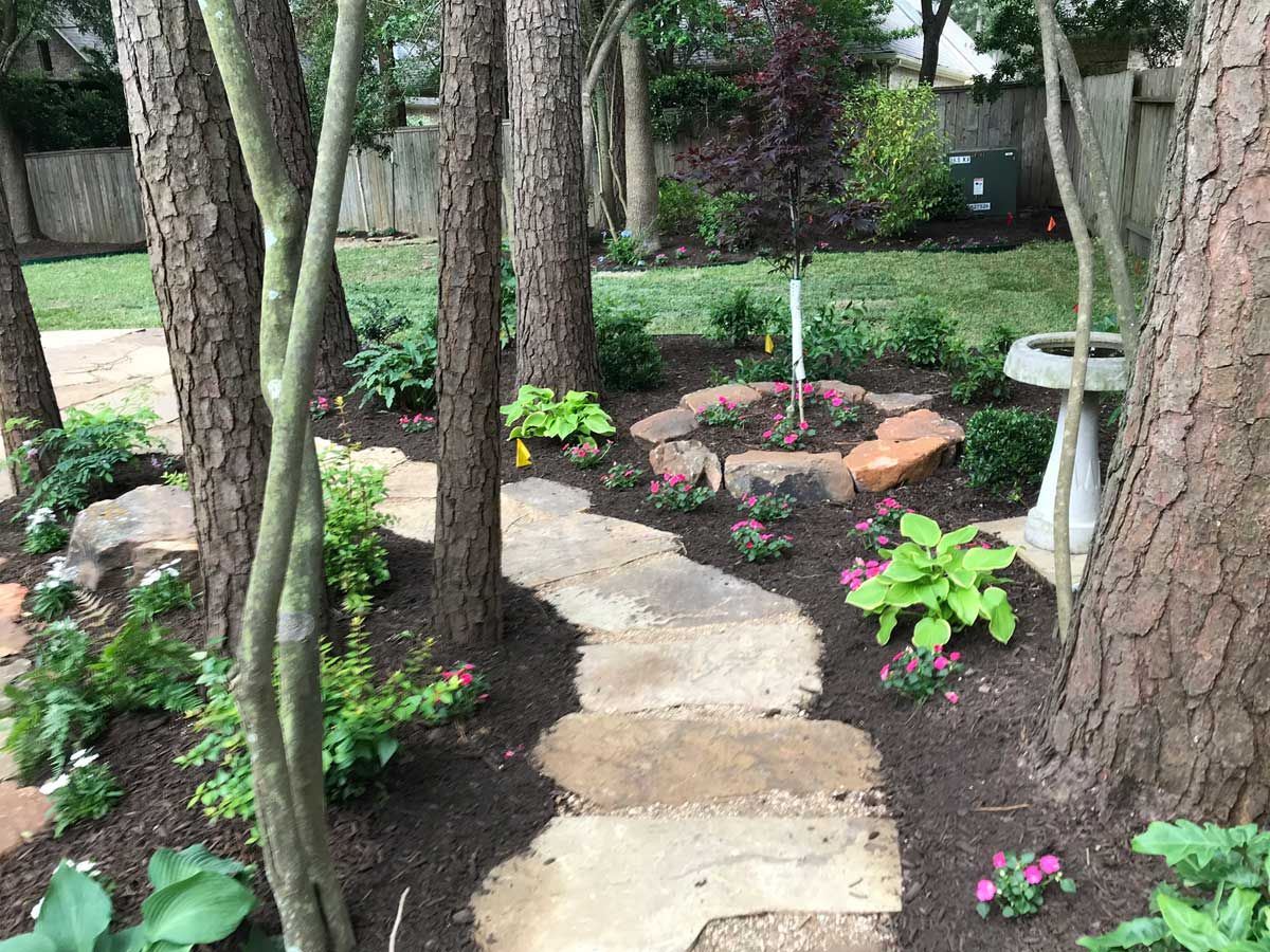 Stone path winds through a garden with trees, flowers, and a bird bath.