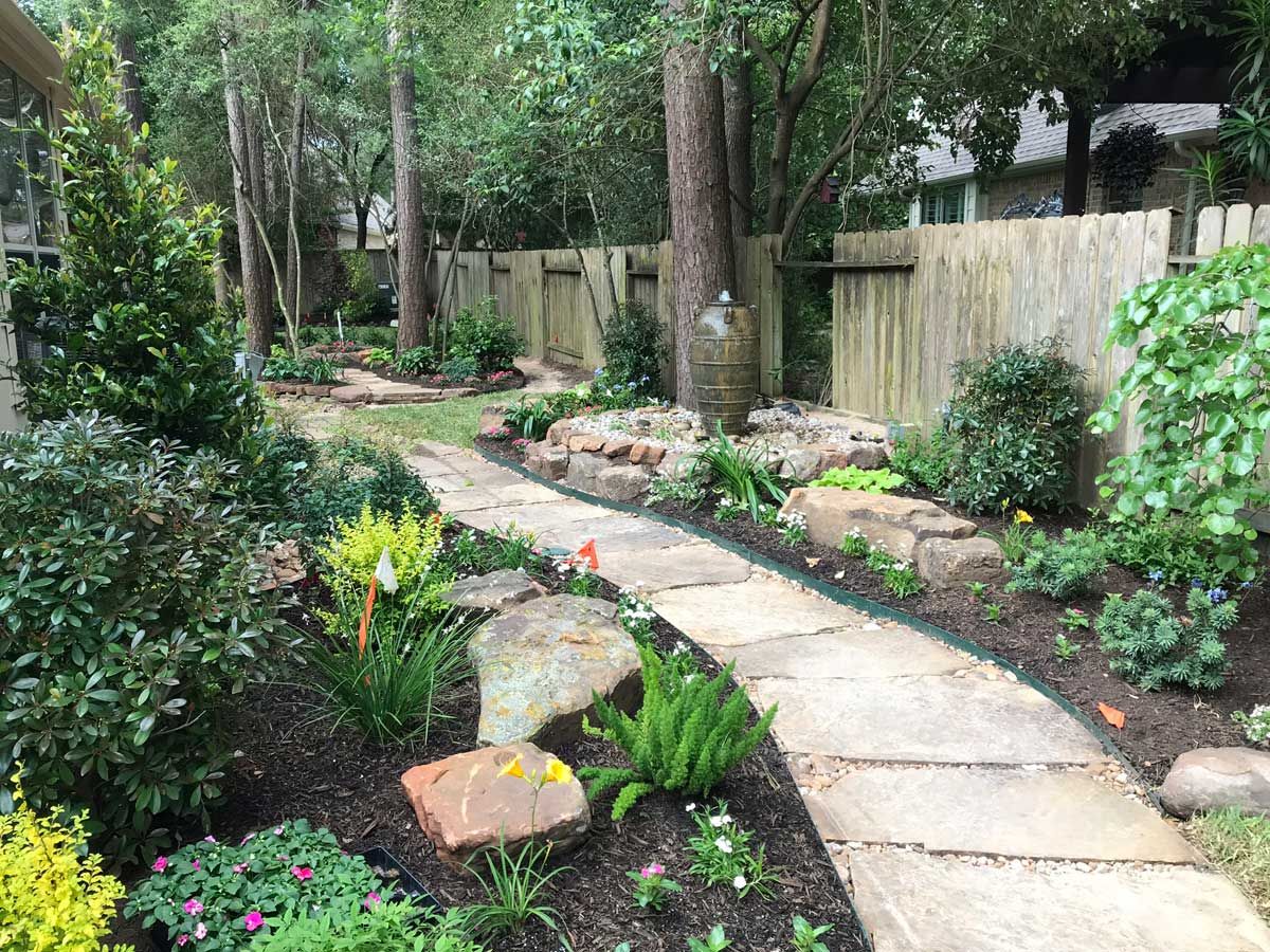 Stone path winds through a landscaped backyard with trees, flowers, and a wooden fence.