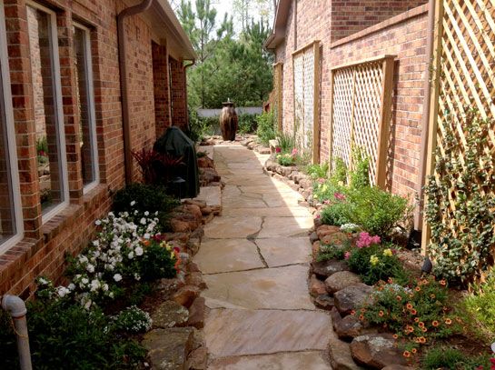 Stone pathway between brick buildings, lined with flowerbeds.