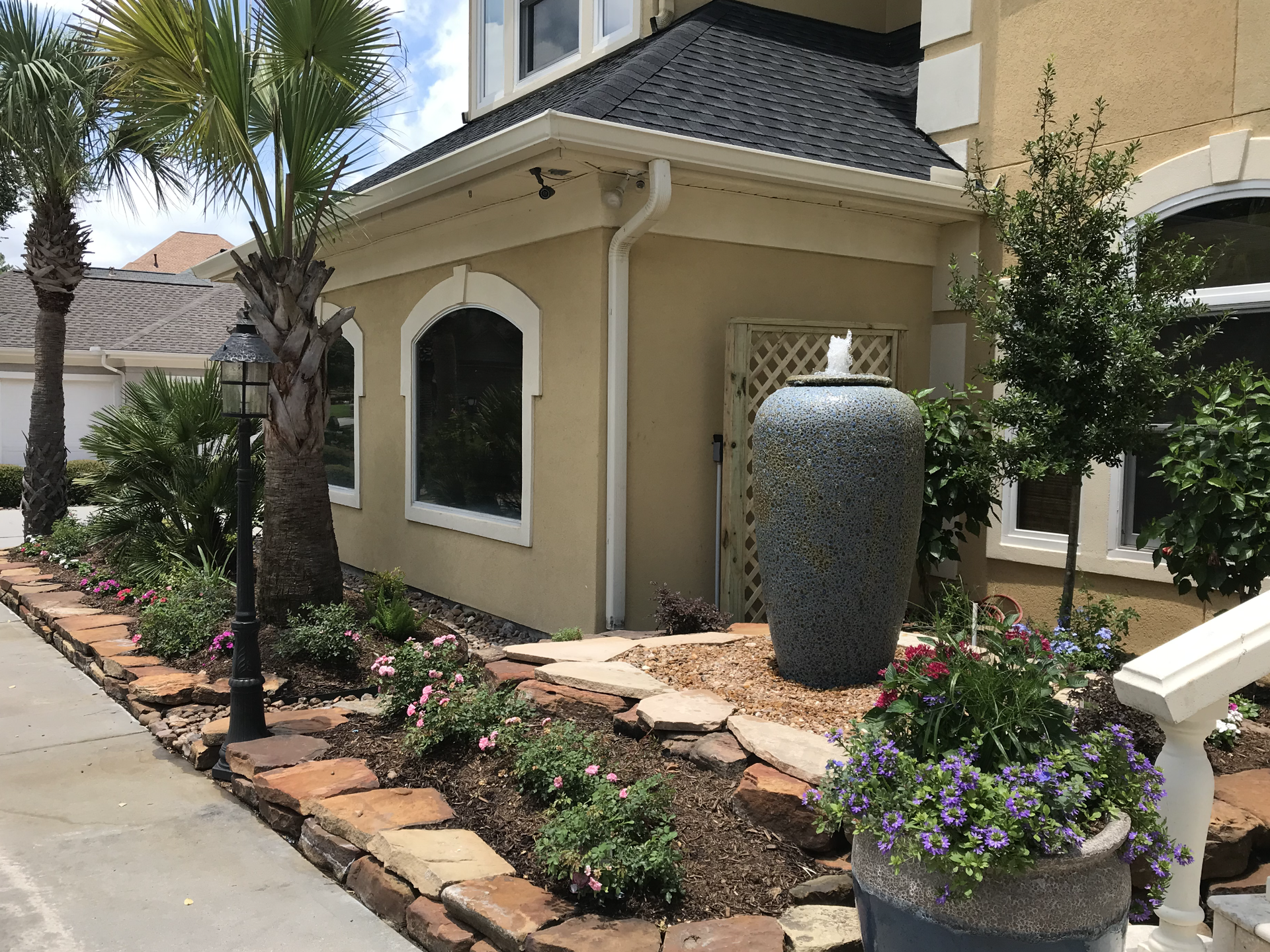 Beige building with stone planter boxes and a large fountain. Palm trees and flowers are present.