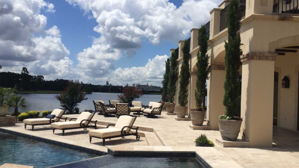 Outdoor patio with lounge chairs and pool overlooking a lake under a cloudy blue sky.