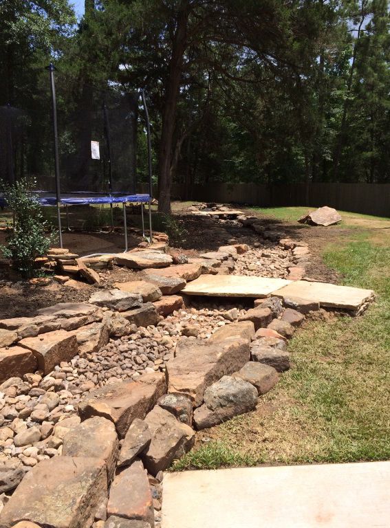 Stone-lined dry creek bed in a backyard with a trampoline, a bridge, and patches of grass.