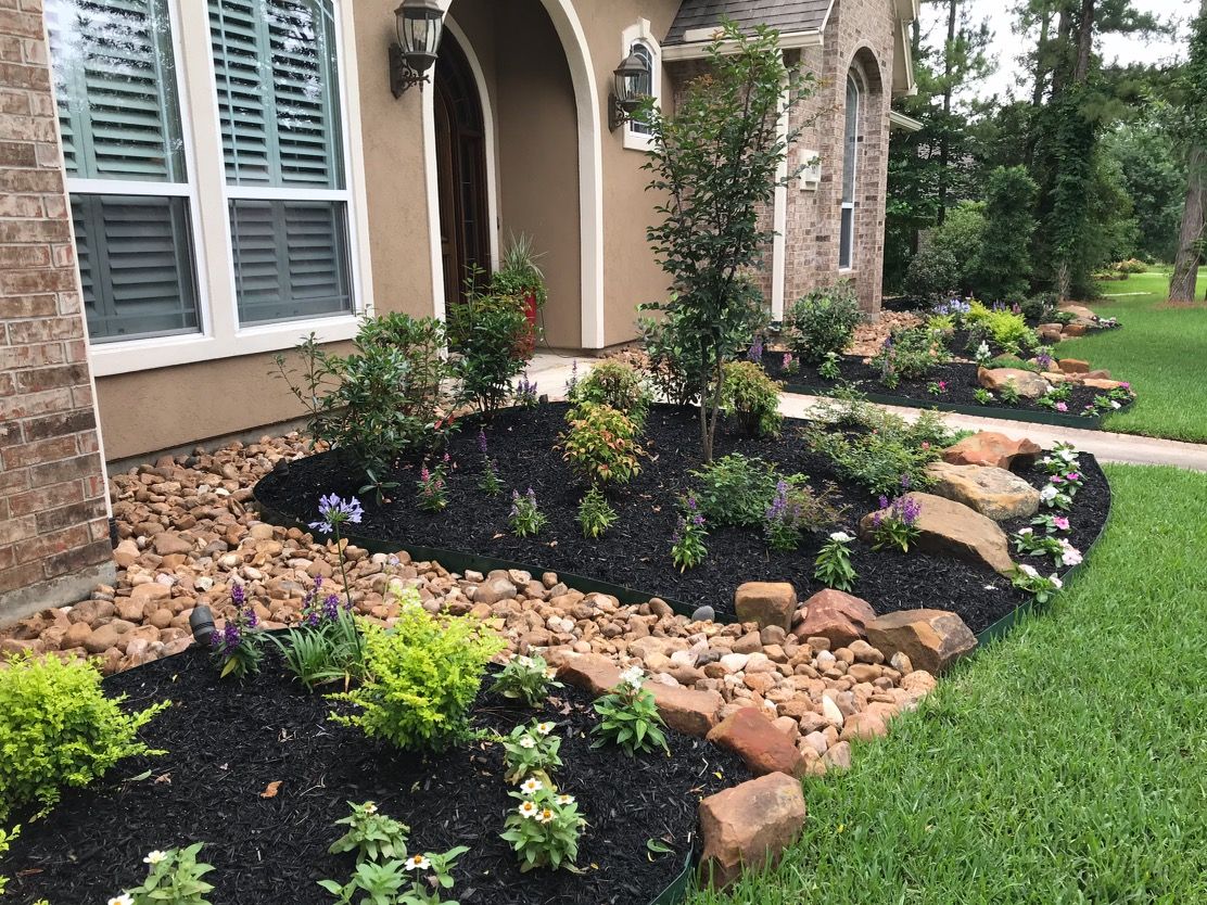 Landscaped front yard with mulch, rocks, and colorful flowers near a house.