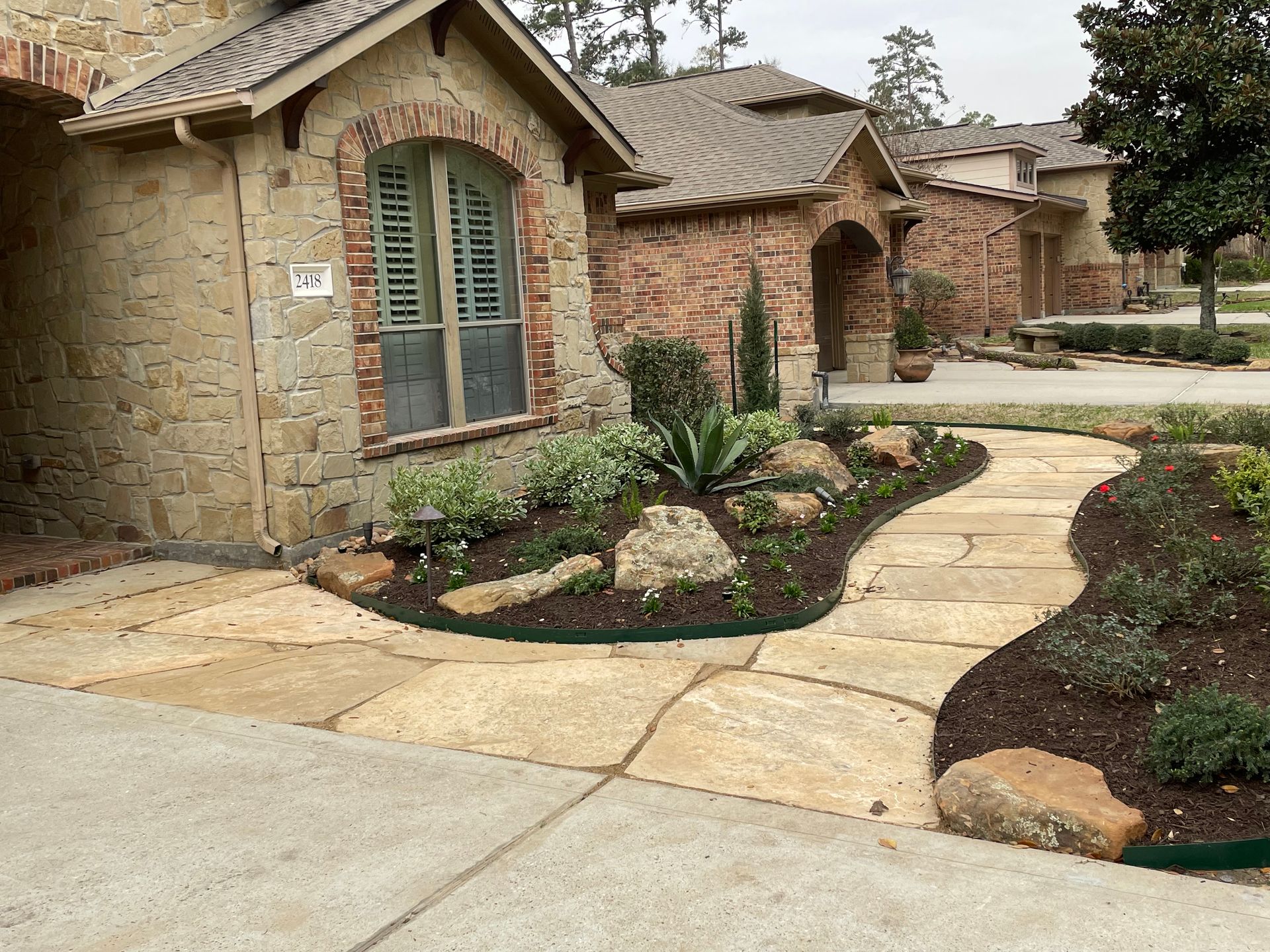 Stone pathway leads to a house with brick and stone facade, surrounded by landscaping and greenery.