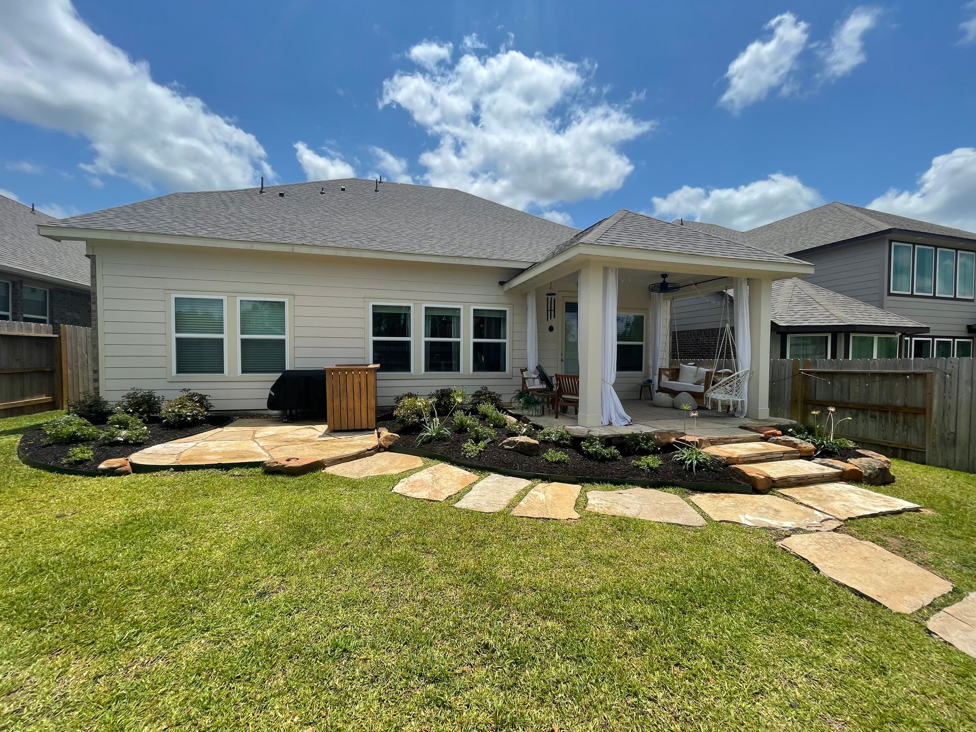 Backyard with patio, stepping stones, flower beds, and a light-colored house under a blue sky.