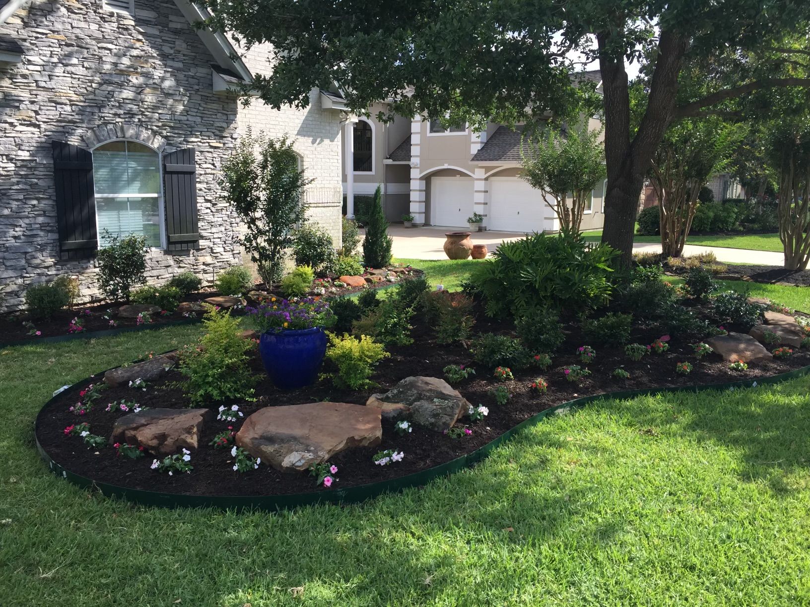 Landscaped front yard with stone house, flower beds, and a tree.