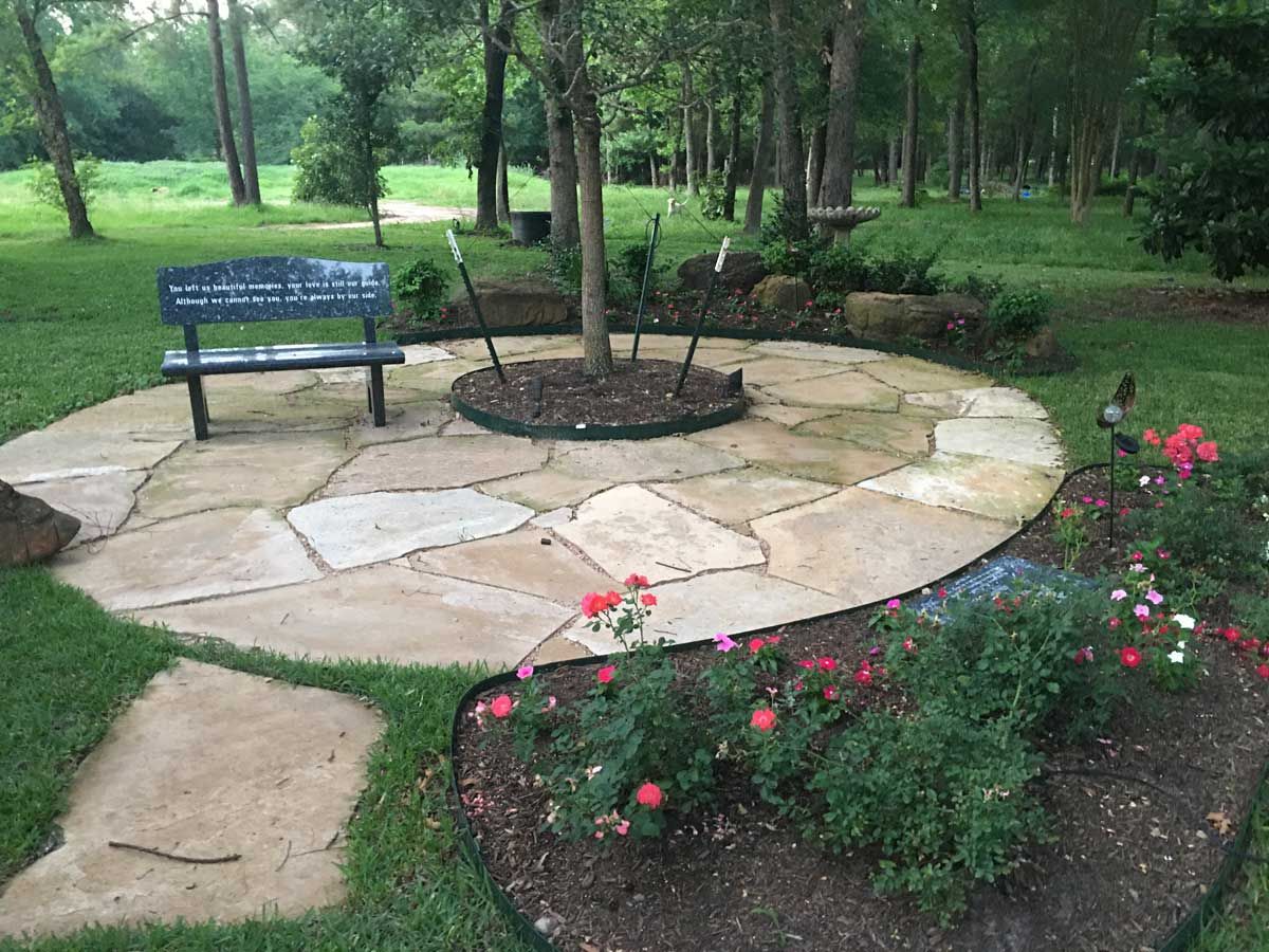 Stone patio with a bench, tree, and flowers in a park-like setting.