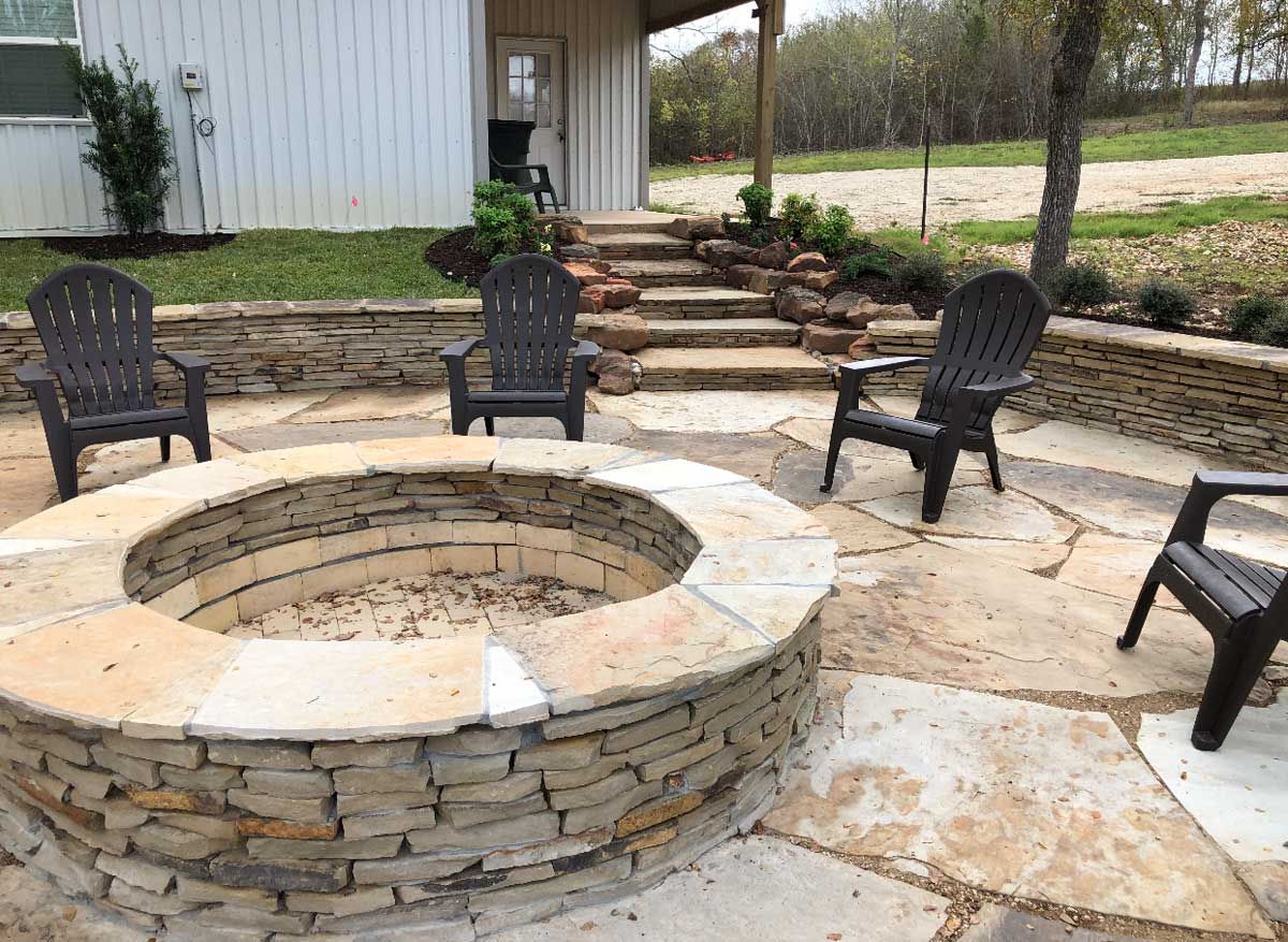 Stone fire pit with four chairs on a patio. Steps lead to a building in the background.