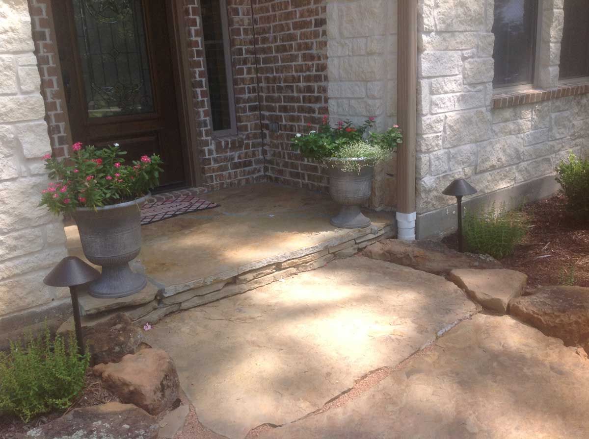 Concrete porch with potted flowers and pathway, lit by garden lights, next to brick and stone facade.