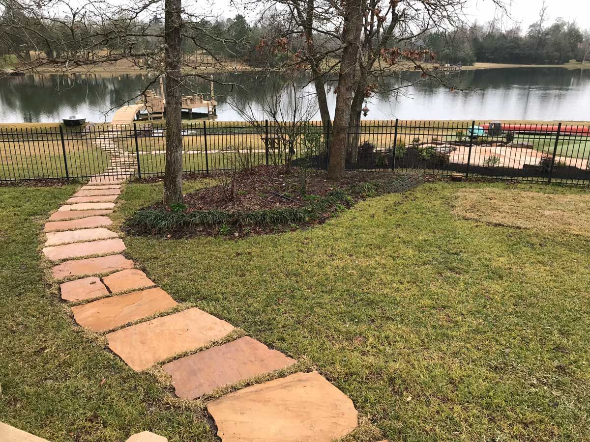 A stone pathway curves toward a lake, surrounded by grass, trees, and a black fence.