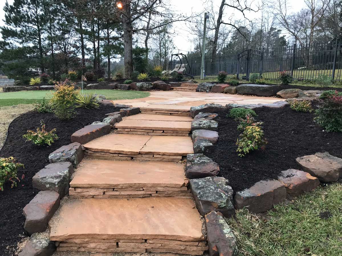 Stone steps lead to a patio surrounded by dark mulch and landscaping, in an outdoor setting.