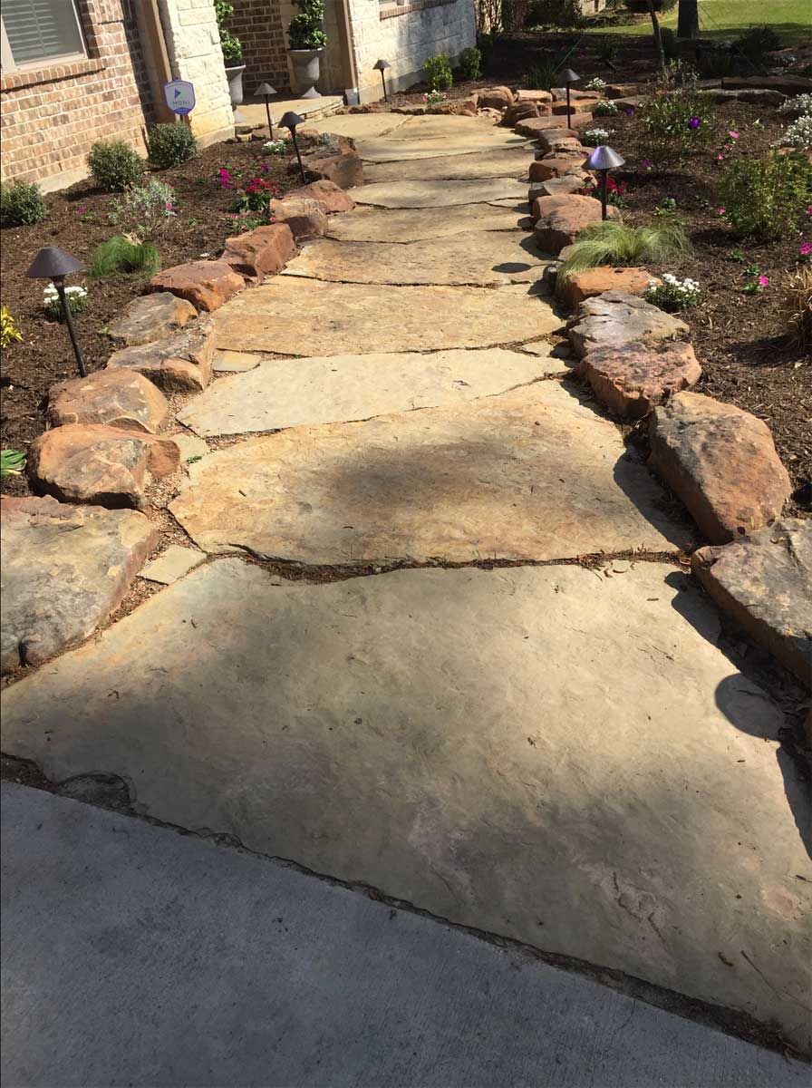 Stone walkway lined with rocks, leading to a house; pathway illuminated by solar lights, surrounded by landscaping.