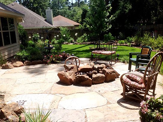 Backyard patio with stone fire pit, seating, and lush landscaping on a sunny day.