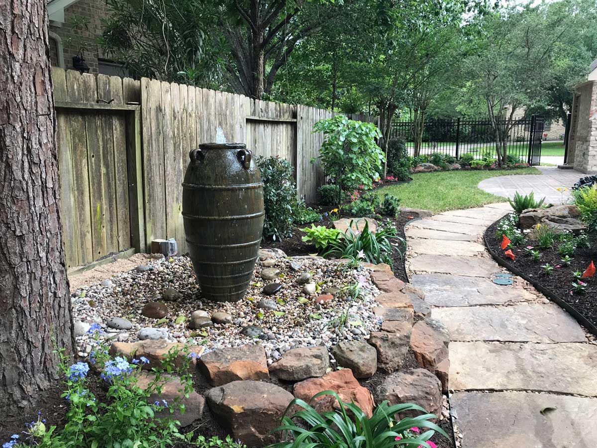 Backyard with fountain, stone pathway, and flower beds near a wooden fence.