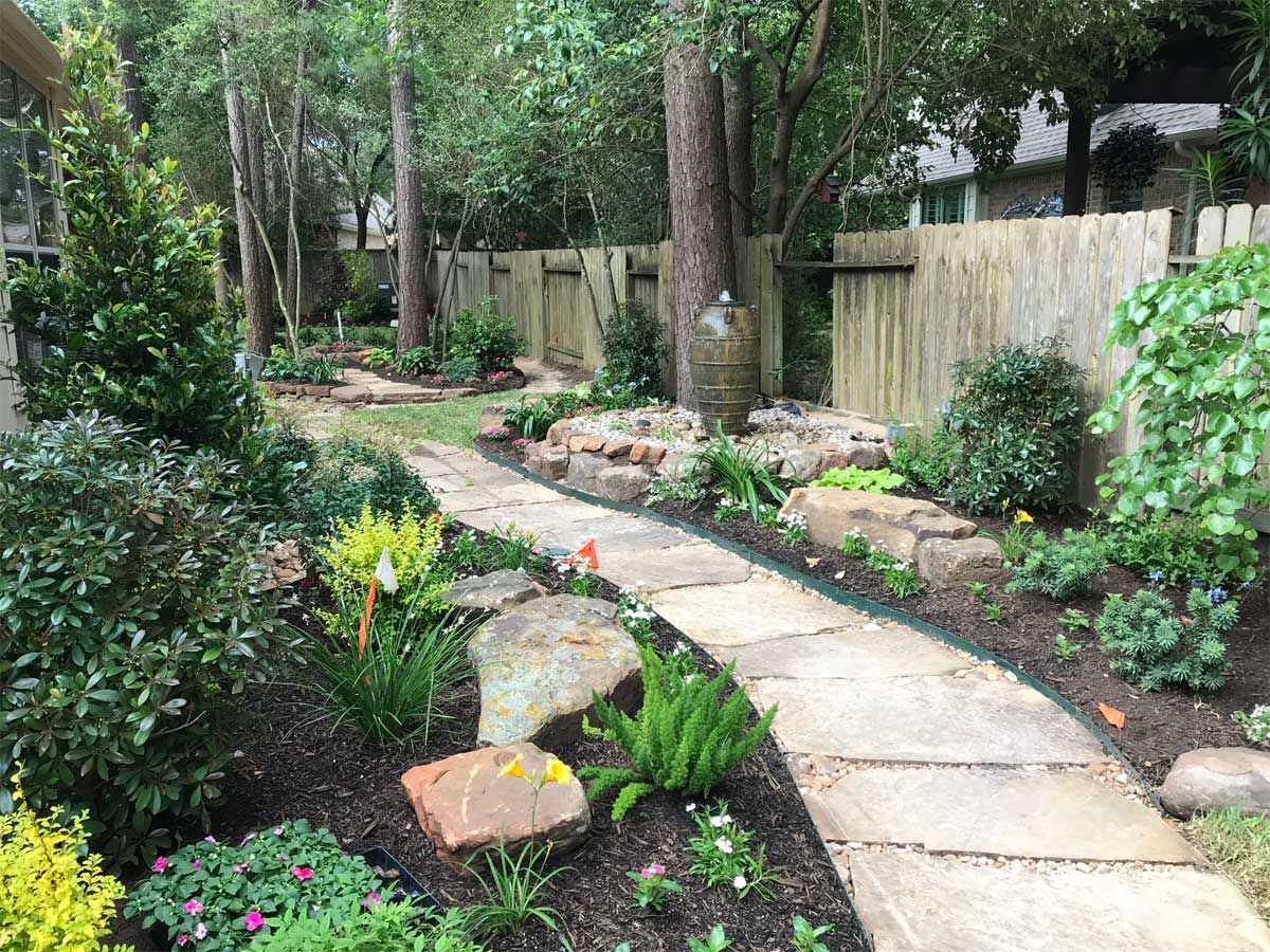 Stone path winds through a landscaped garden with trees, shrubs, and a wooden fence in the background.