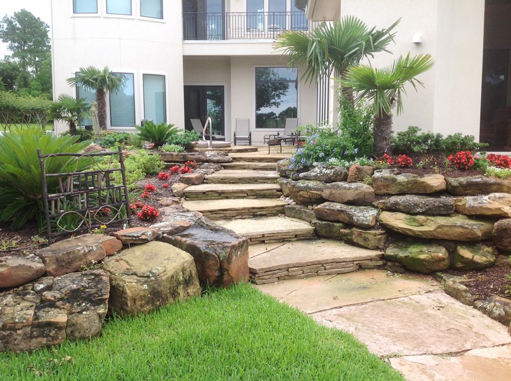 Stone steps leading up to a two-story house, lined with rock gardens and palm trees.