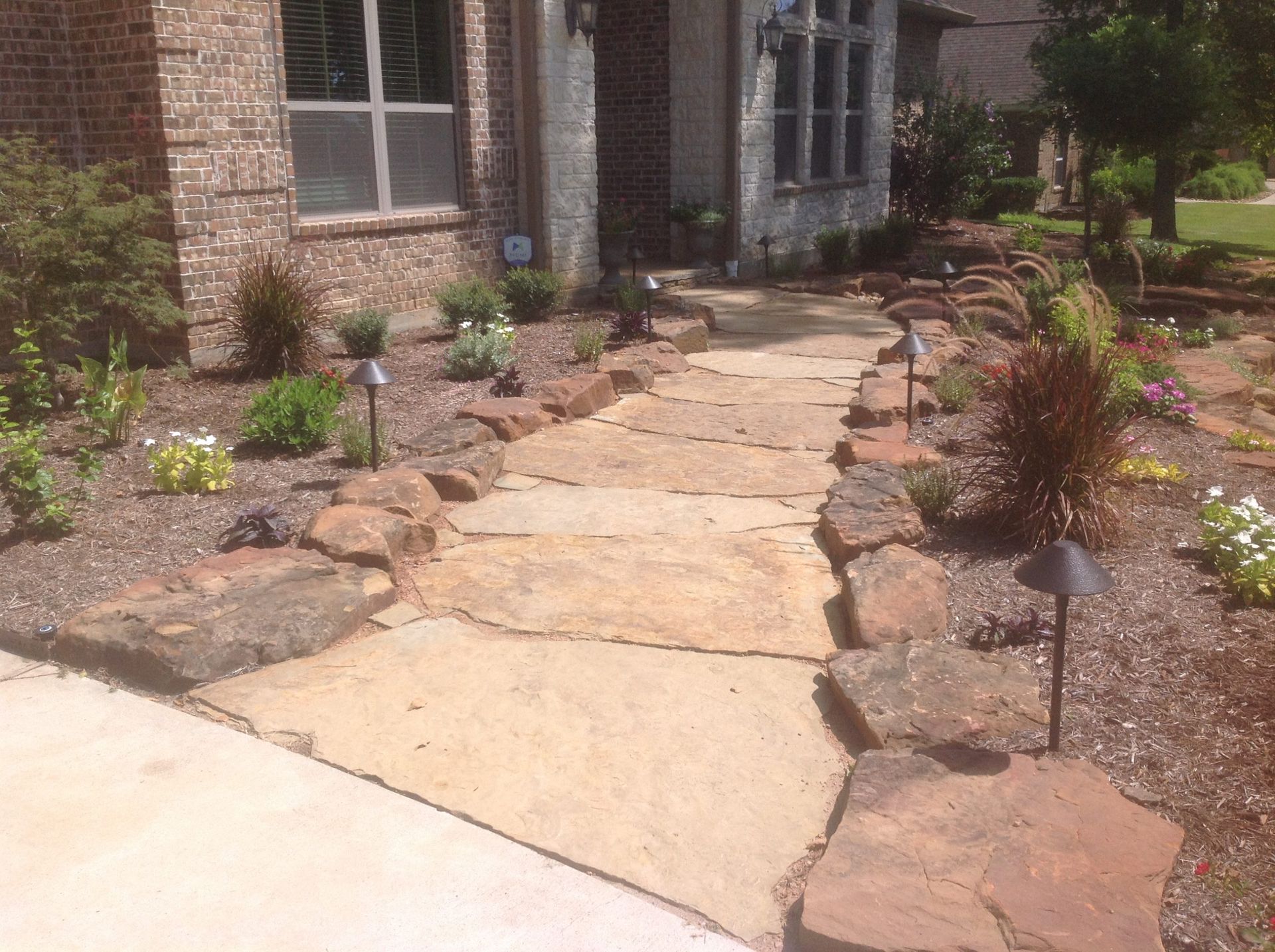 Stone pathway leading to a house, bordered by landscaping with brown mulch and lighting fixtures.