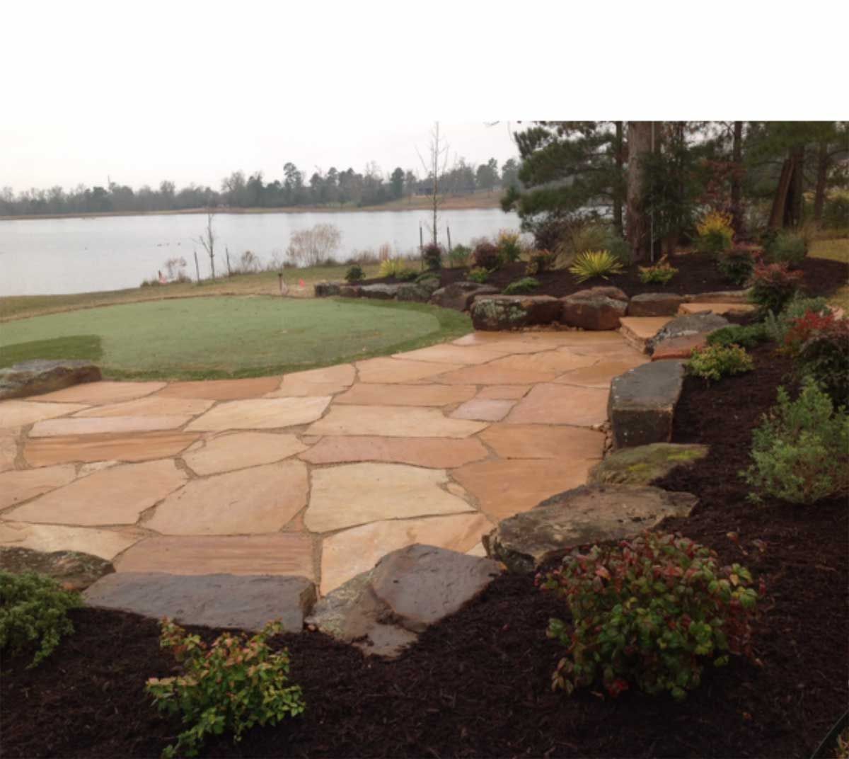 Stone patio next to putting green and lake, surrounded by plants and mulch.