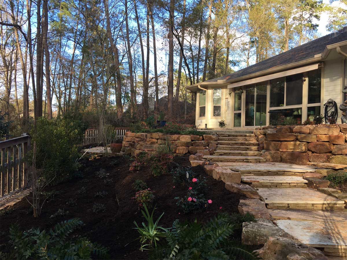 Stone steps lead up to a house, with a stone retaining wall and garden in a wooded setting.