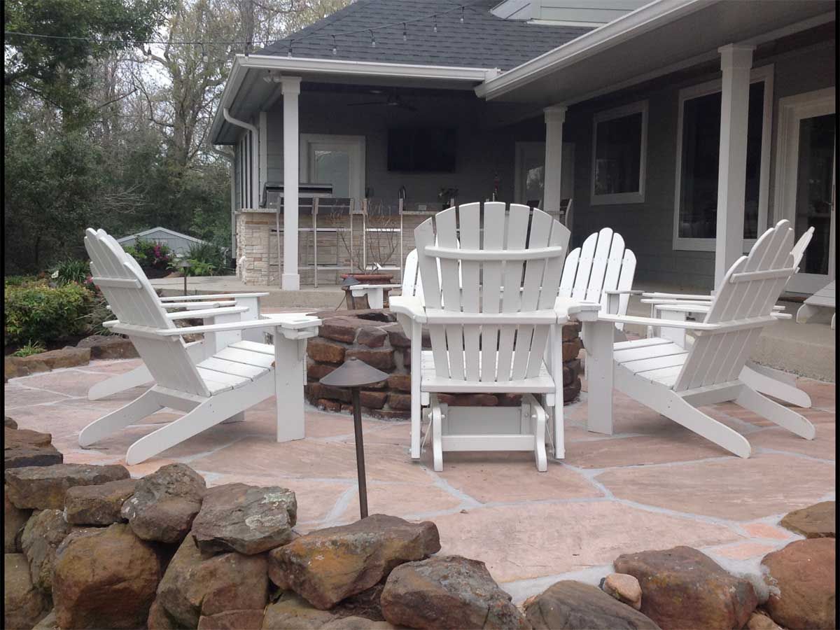 White Adirondack chairs around a fire pit on a stone patio, near a house with a porch.