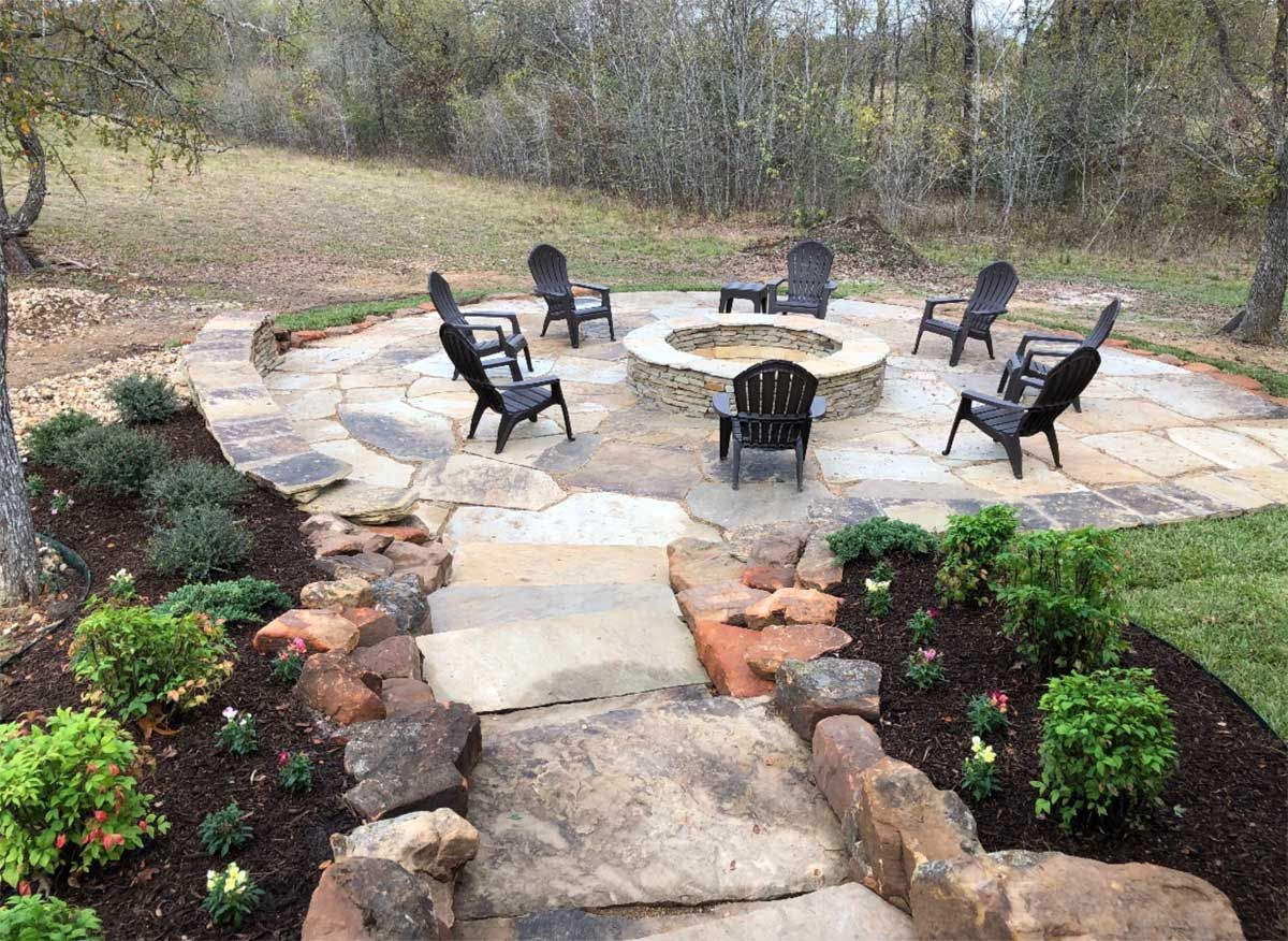 Stone fire pit surrounded by black chairs, on a stone patio, with landscaped path.