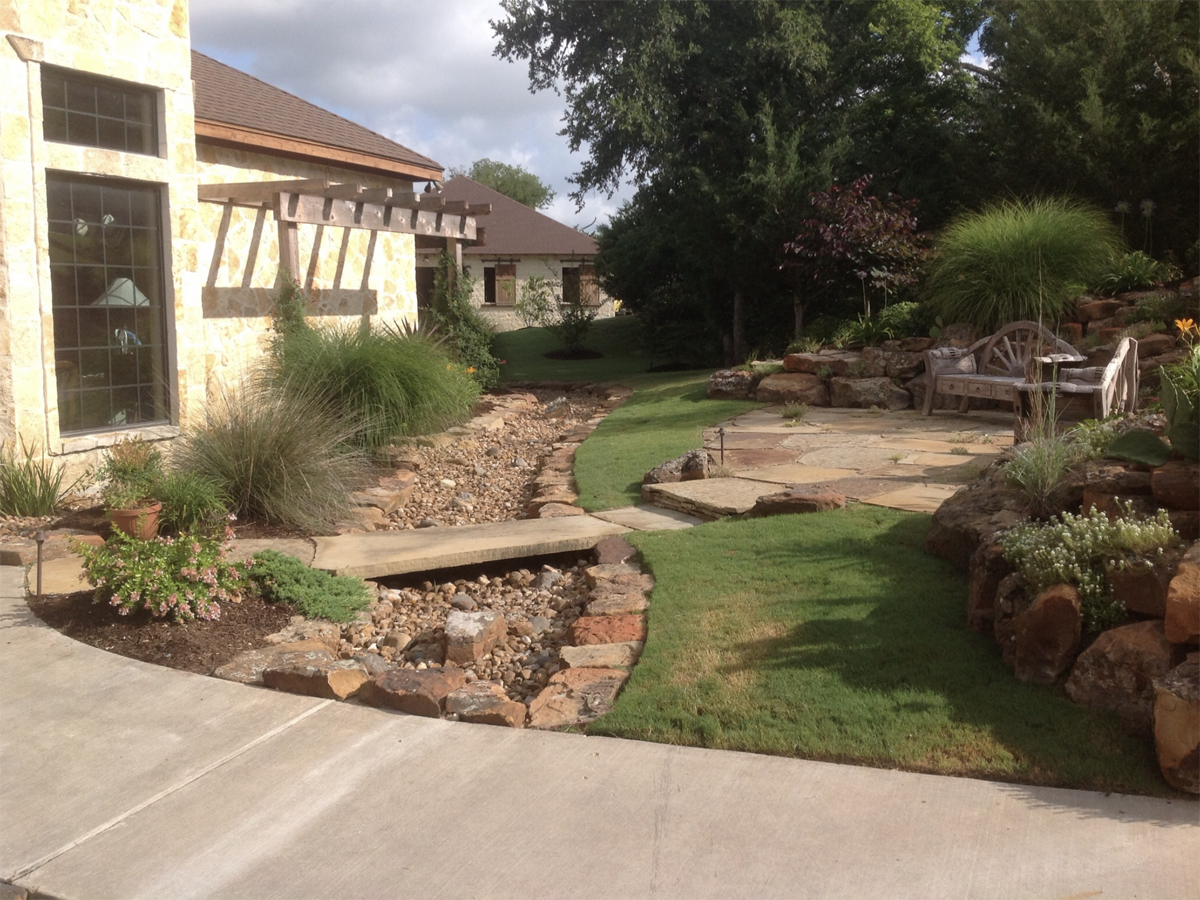 Stone path and bridge in a landscaped yard with a house in the background and a seating area.