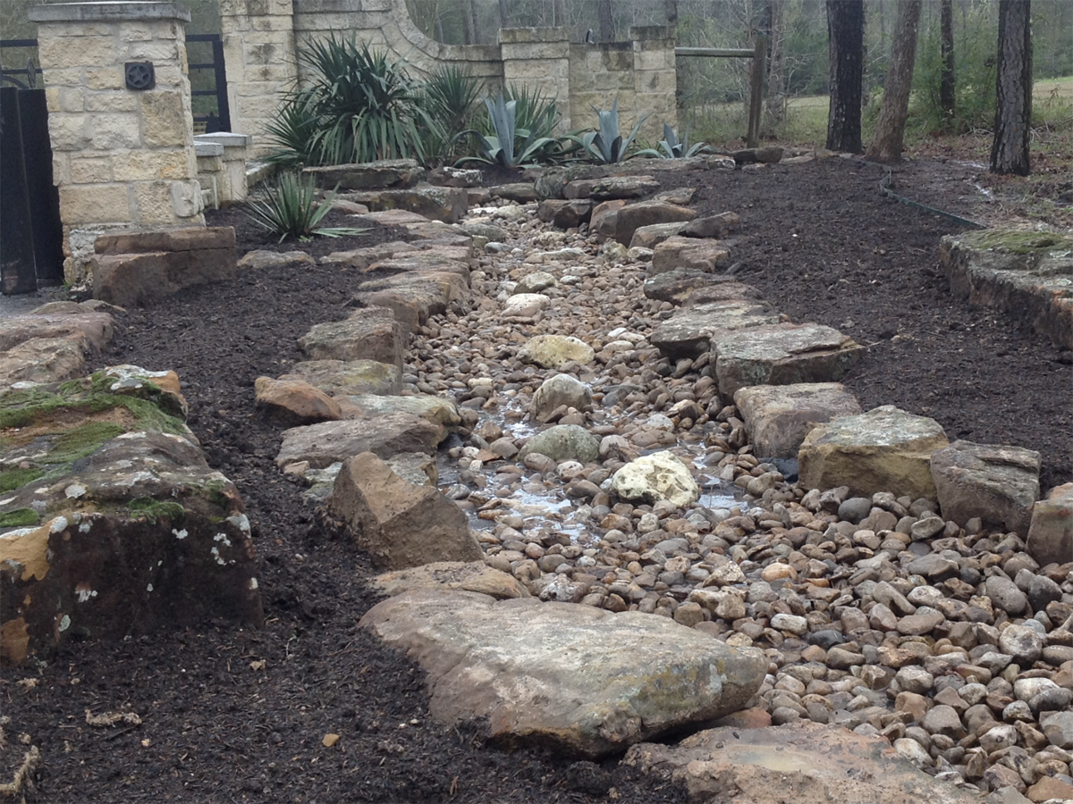 Stone-lined water feature with river rock and mulch in a garden setting.