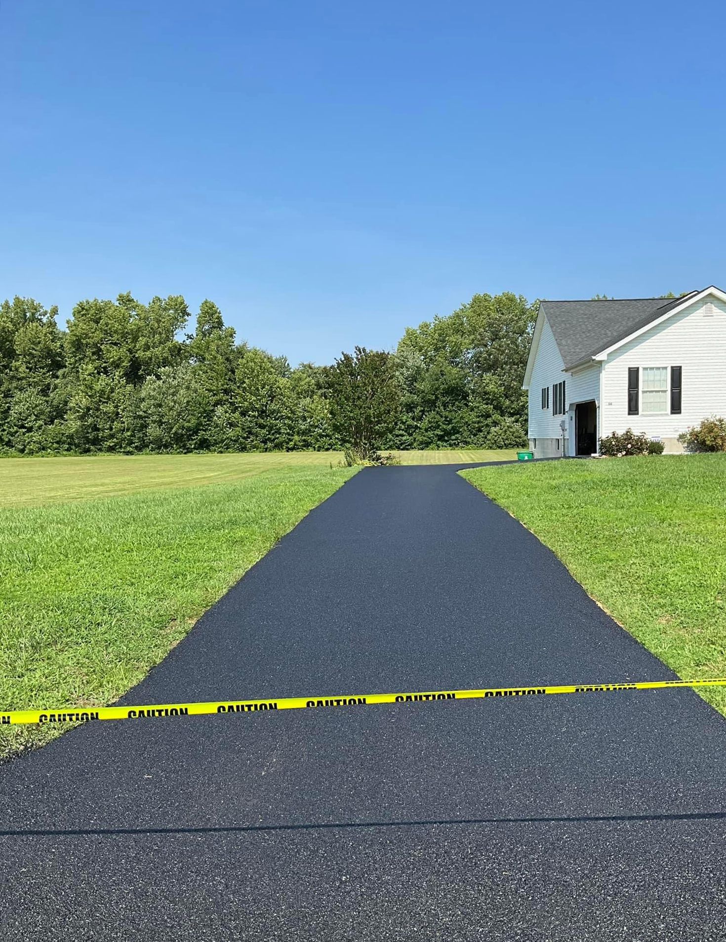 A black driveway leading to a white house in the middle of a grassy field.
