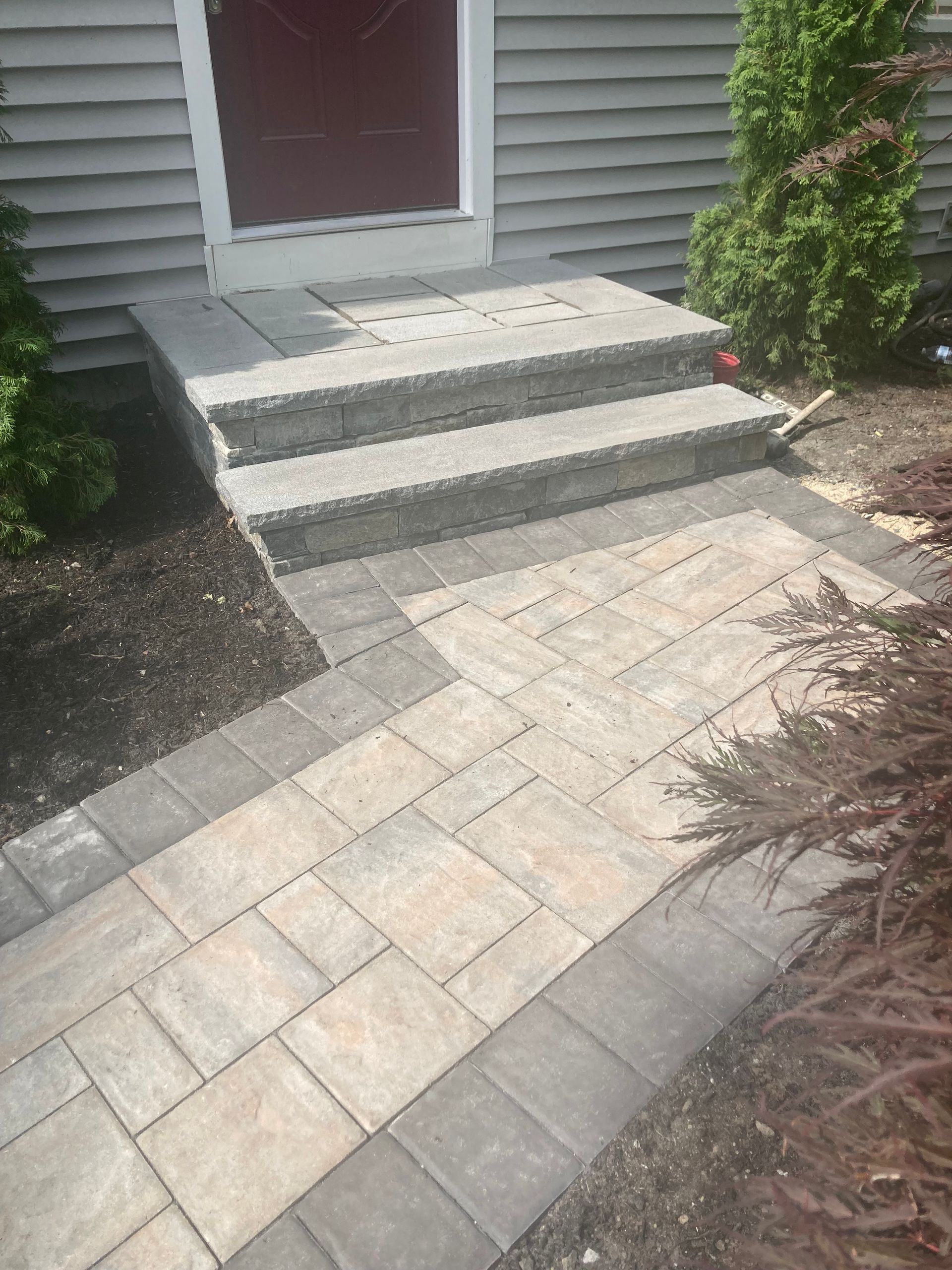 Paver walkway and steps leading to a front door. Gray and tan stones, brown trim, bushes on either side.