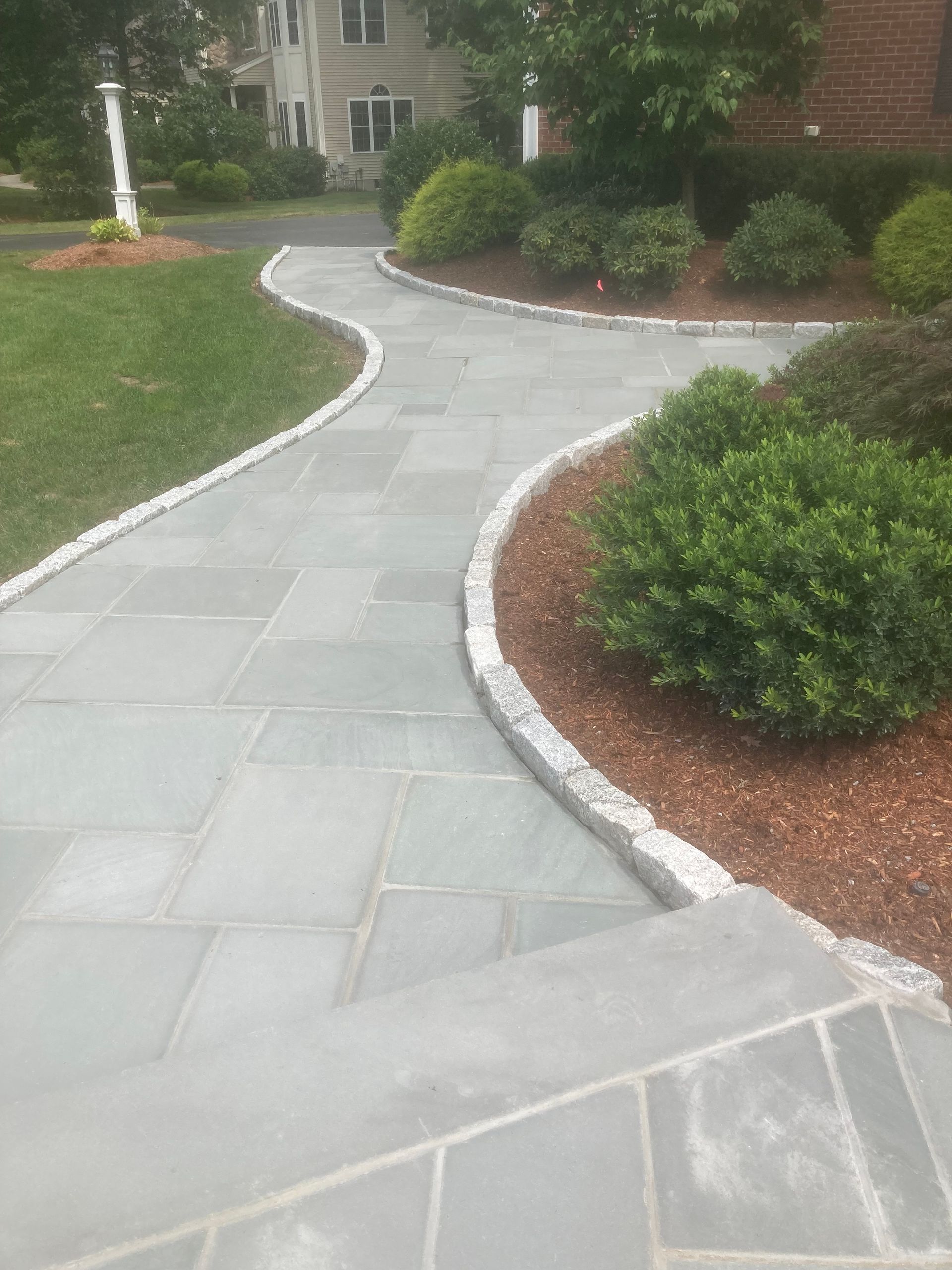 Stone walkway winding through landscaped yard, bordered by bushes and mulch.