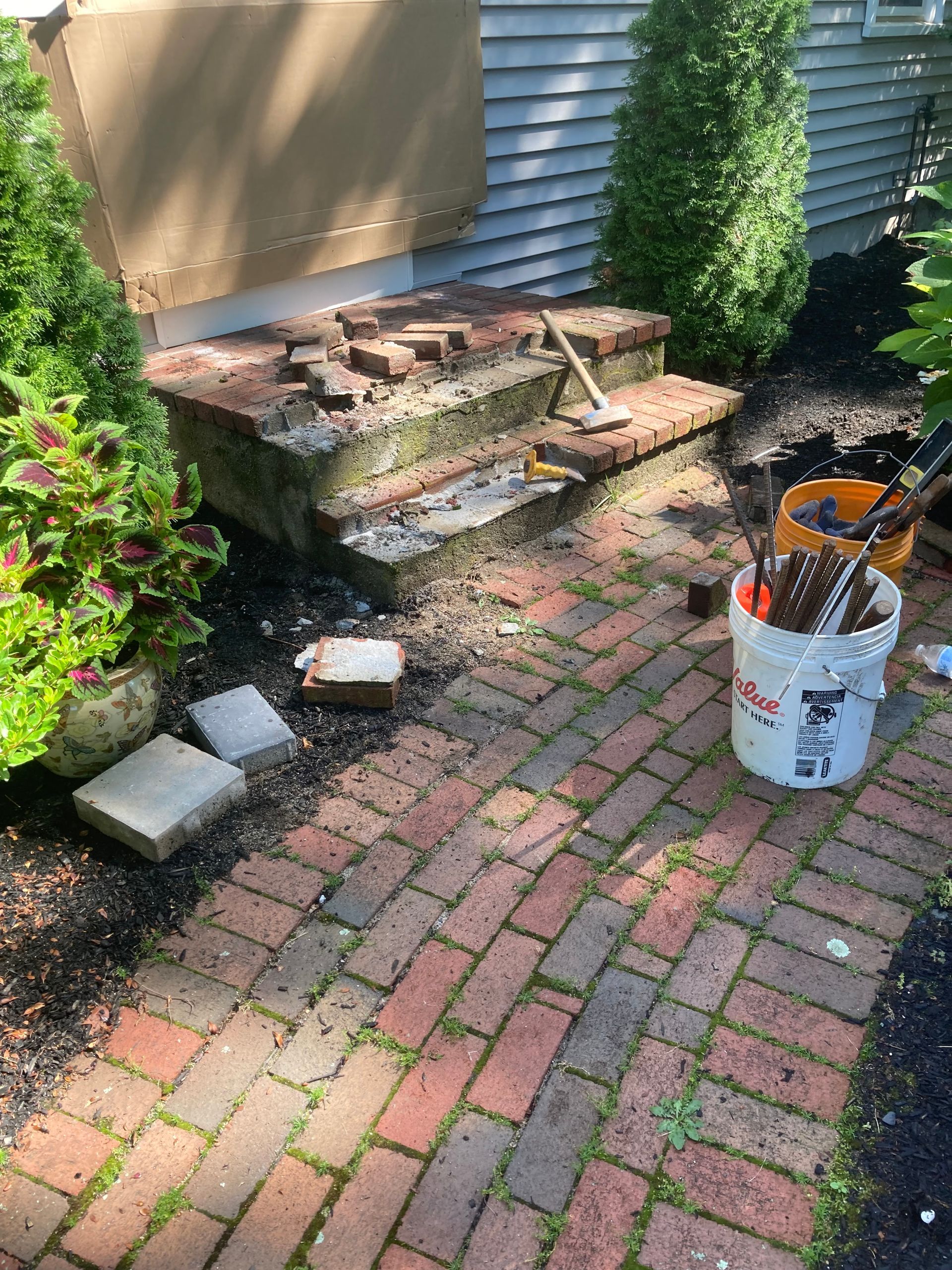 Damaged brick steps being repaired, with tools nearby, set in a brick patio with overgrown weeds.