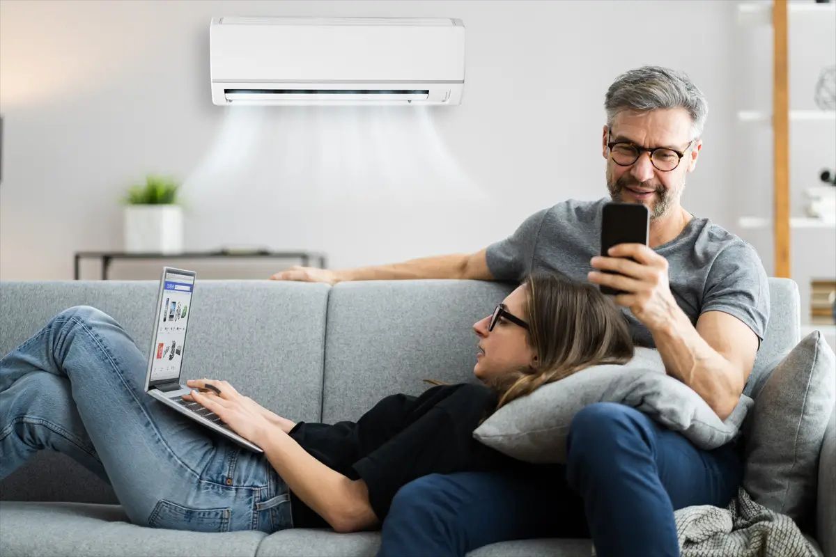 Couple relaxing on a couch under an air conditioner, one using a laptop, the other a phone.