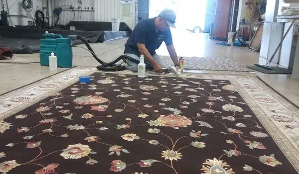 Worker cleaning a large floral rug in a workshop with a floor buffer.