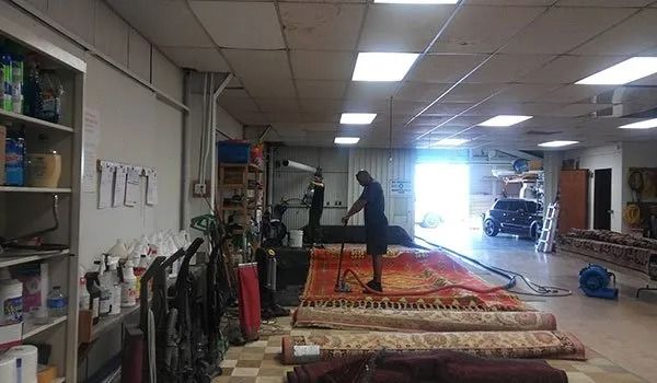 Indoor workshop with rugs laid out, a person standing on a central carpet, shelves and bright window in back