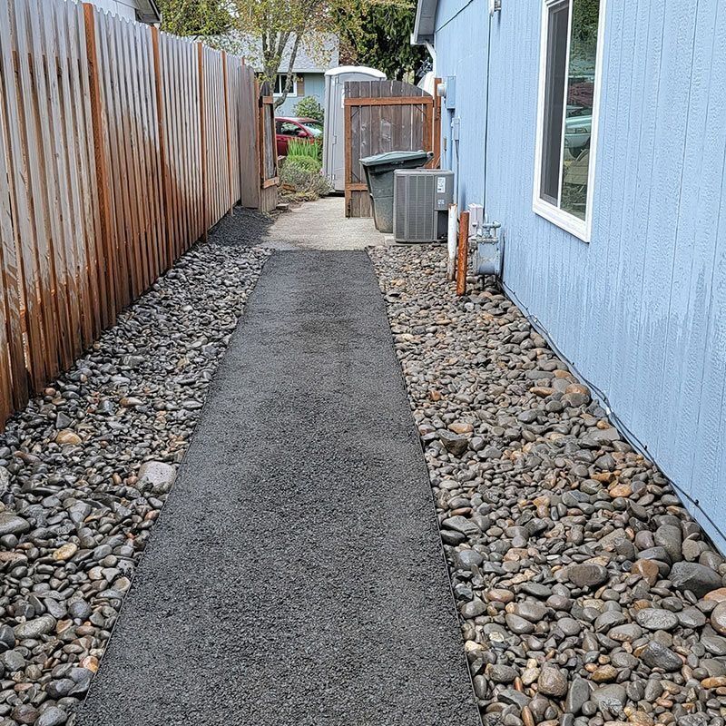 A narrow path with a dark asphalt walkway lined by gray gravel stones between a wooden fence and a light blue wall.