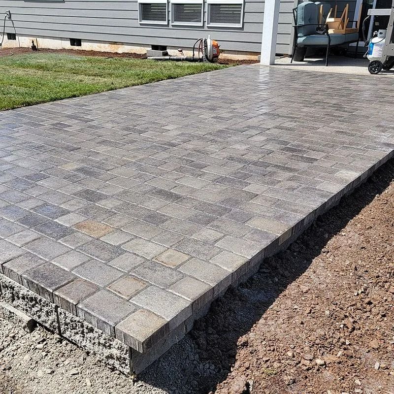 A newly installed gray paver patio bordering a house foundation with grass and a dirt area nearby.
