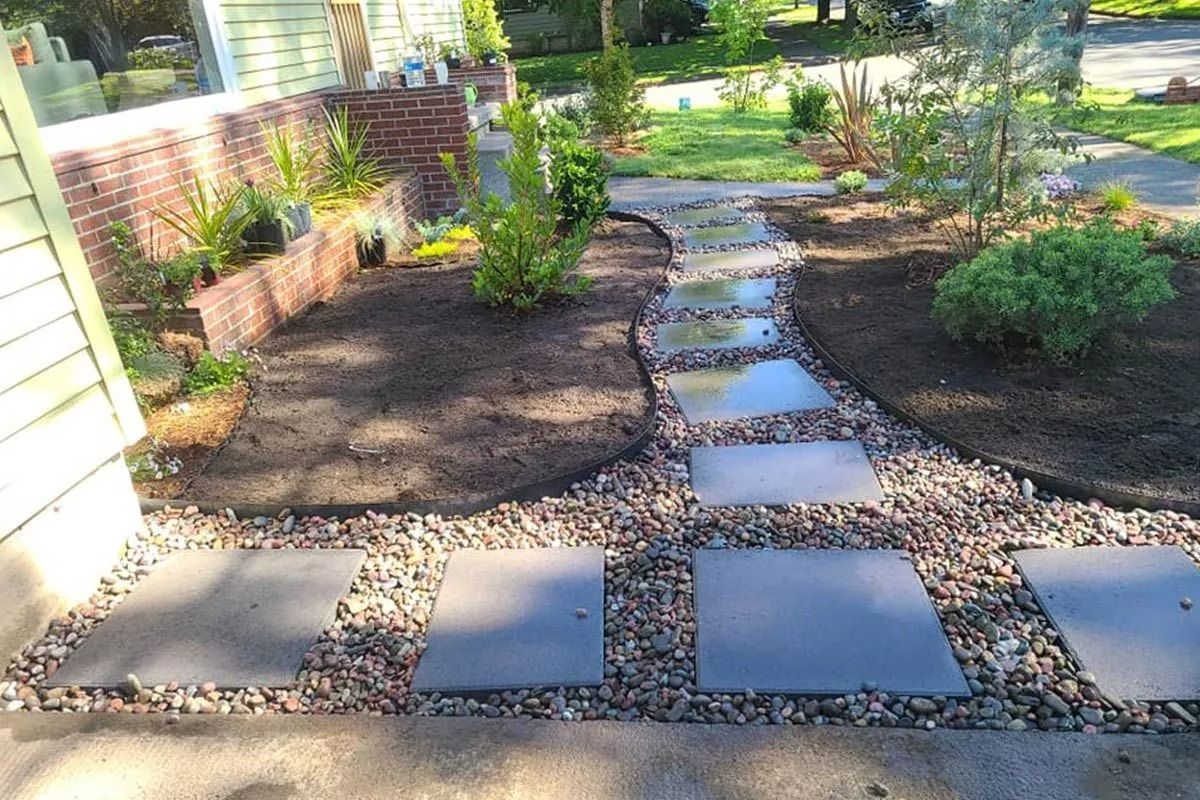 A stone paver walkway winds through a landscaped front yard with dark mulch and green plants next to a brick house.