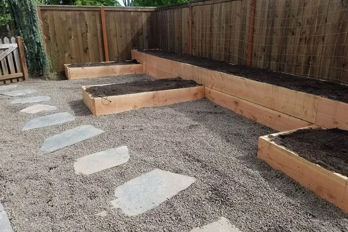 Tiered wooden raised garden beds filled with soil, set against a wooden fence with a gravel path and stone pavers.