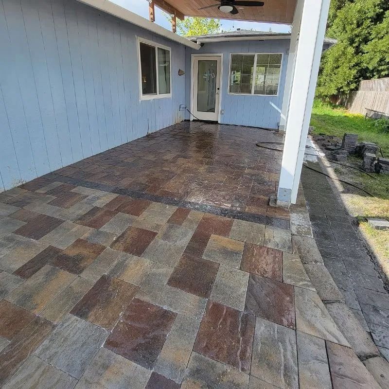 A patio made of multicolored square pavers next to a light blue house with a white door and windows under a wooden porch.