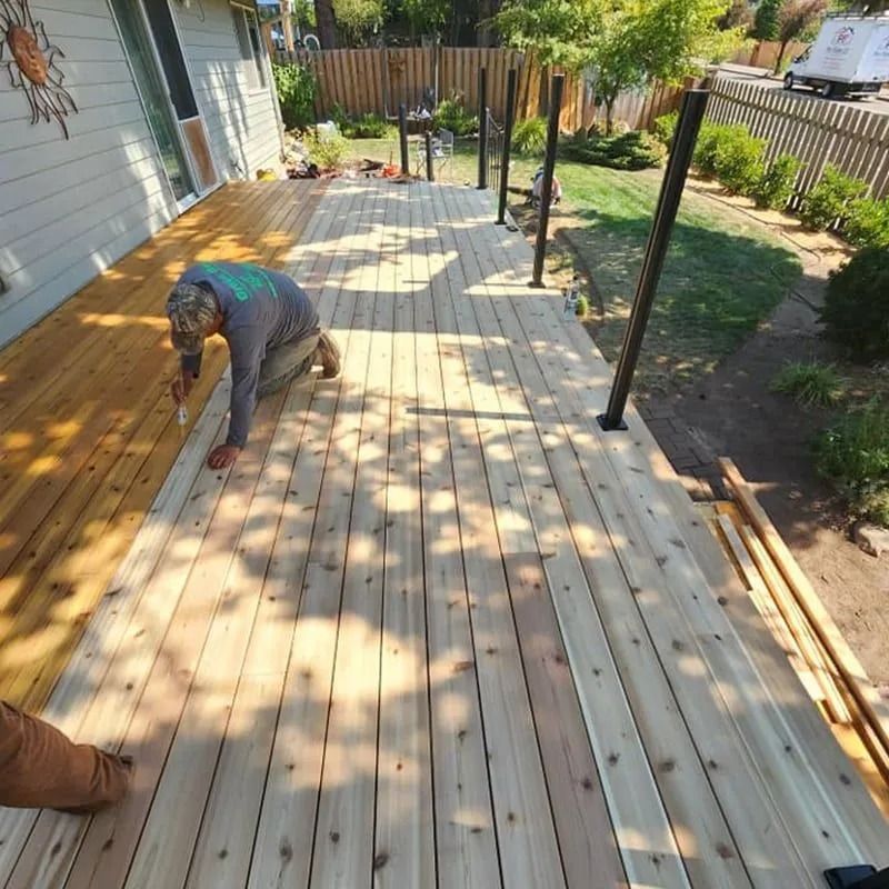 A person kneeling on a wooden deck, applying stain to the boards next to an unfinished, lighter-colored section.