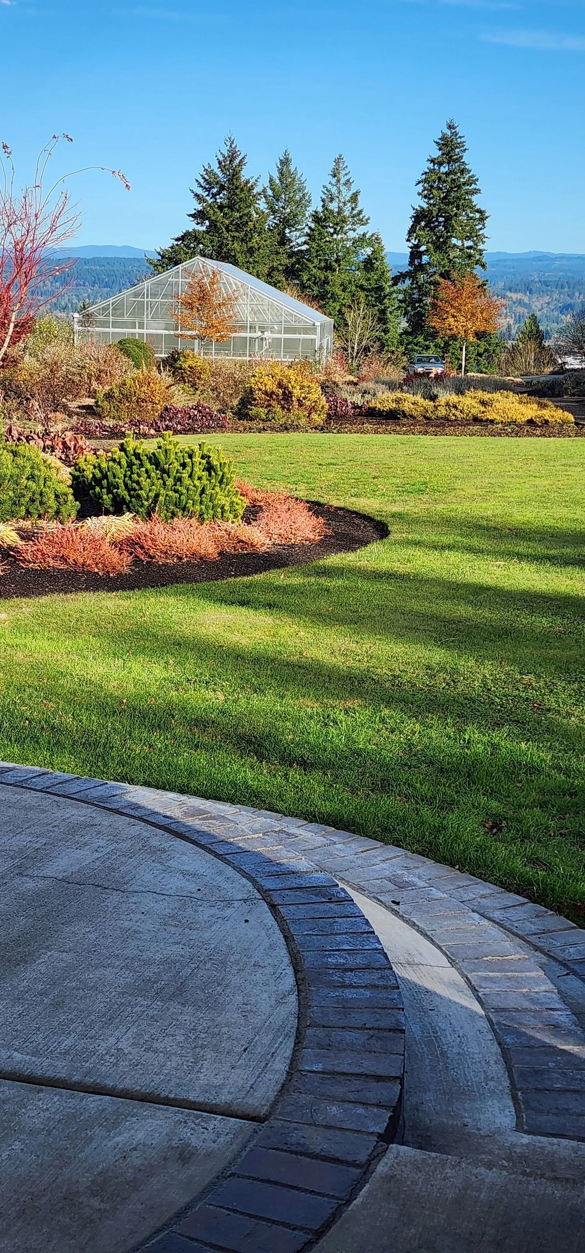 A paved circular patio leads to a lush green lawn, tiered landscaping, a greenhouse, and distant mountains under blue sky.