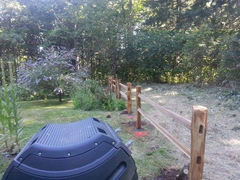 A black compost bin sits in the foreground next to a newly installed wooden split-rail fence in a grassy yard.