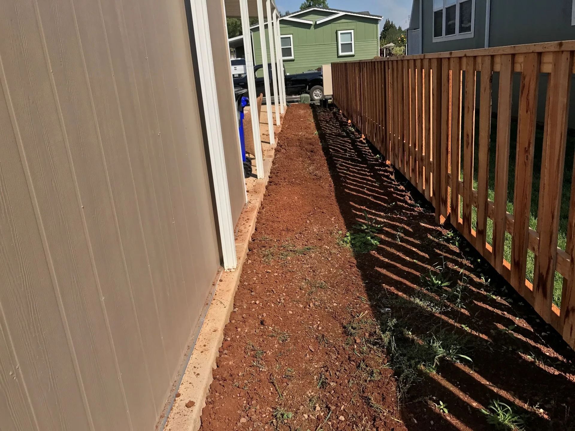 A narrow dirt path between a light beige house wall and a wooden picket fence on a sunny day.