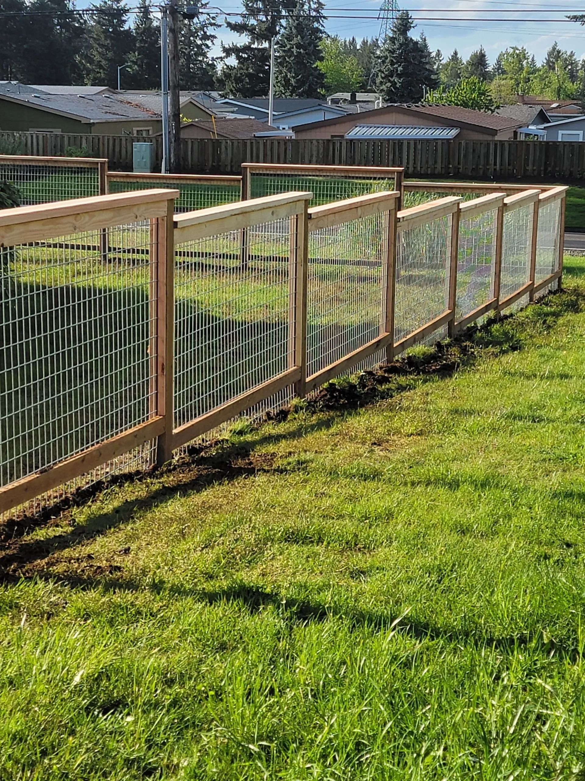 A wooden garden fence with wire mesh panels sits on a grassy lawn under a sunny sky.