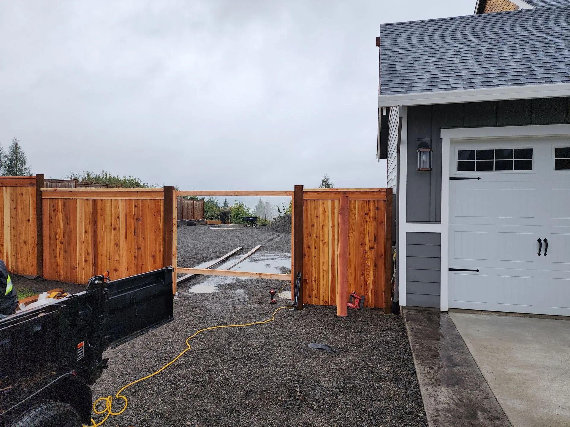 A cedar fence with an open gate frame sits between a house garage and another fence section on a gravel lot.