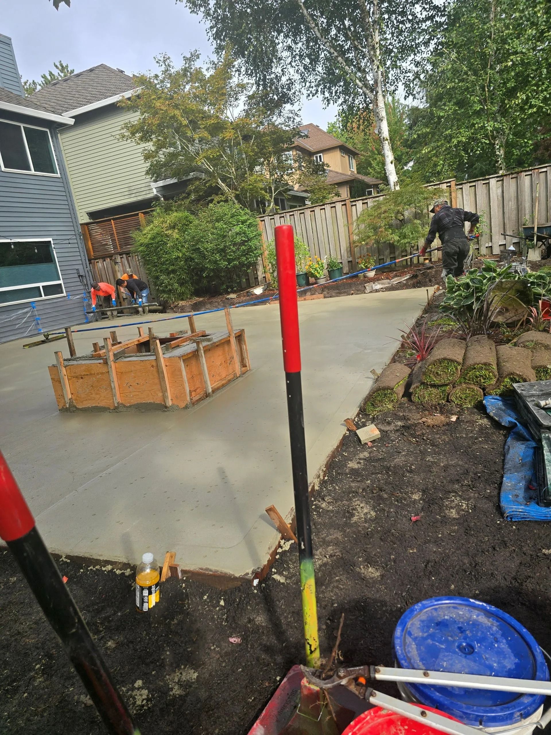 Workers pour concrete for a new patio in a residential backyard, featuring a wooden form for a fire pit in the center.